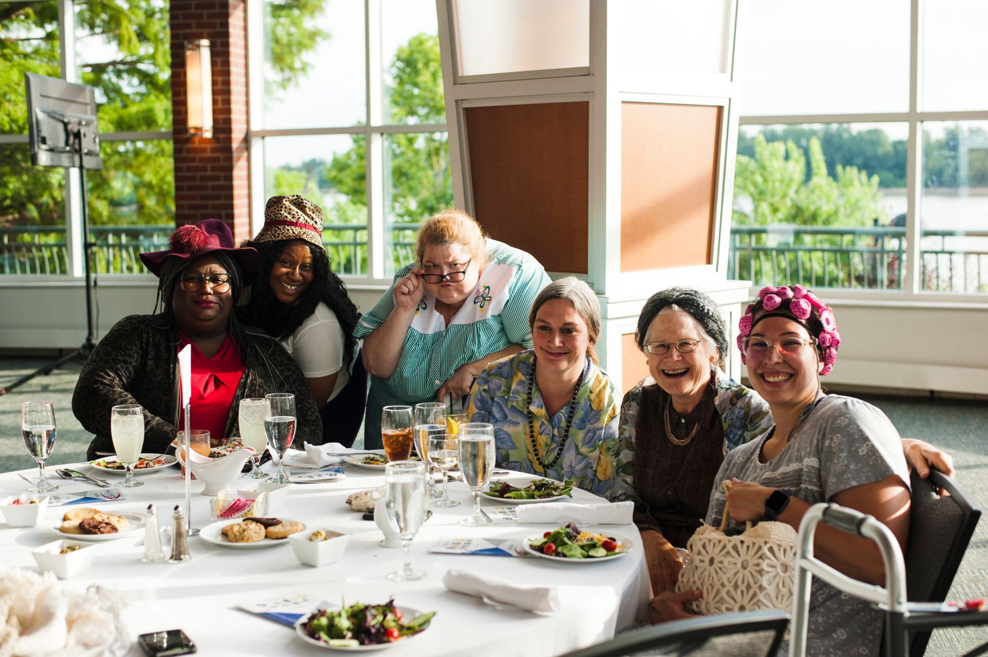 A group of six people sit around a table at a brightly lit, windowed venue, smiling while dining at a catered event.