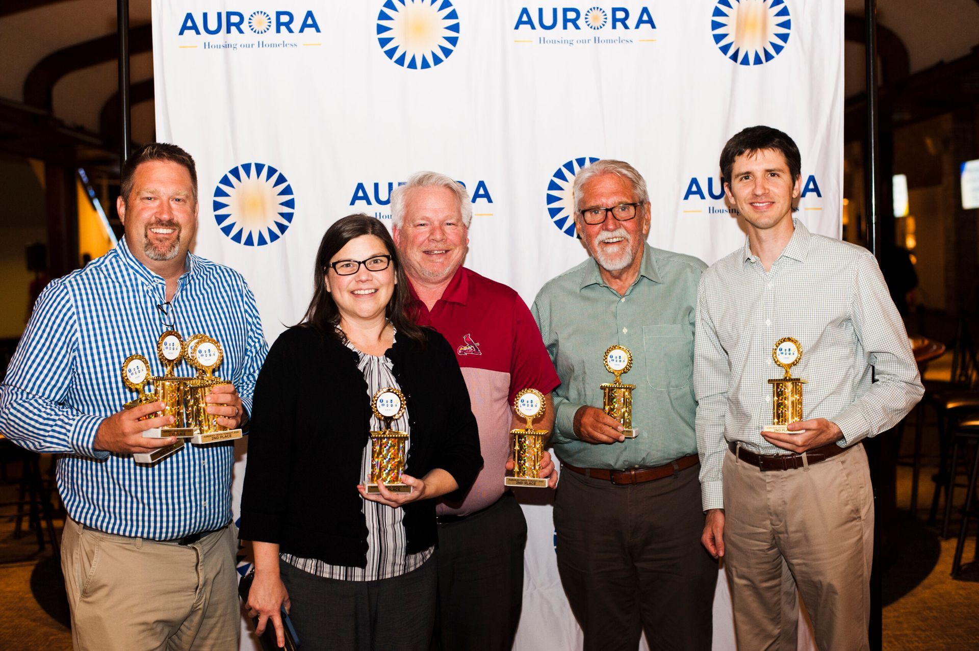 Five people pose holding trophies in front of an Aurora branded backdrop at an indoor event.