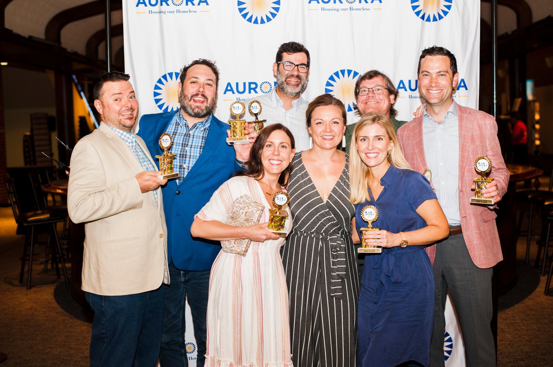 Eight people smiling while holding award trophies in front of an Aurora-branded backdrop at an evening event.