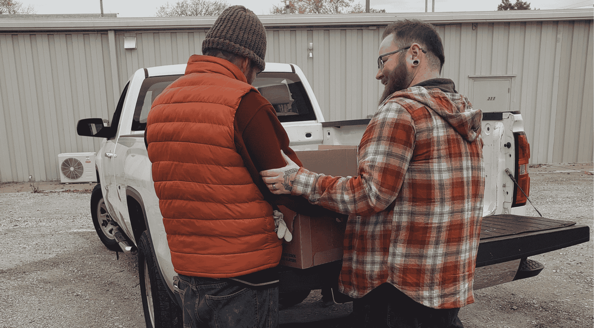 Two individuals in casual outdoor clothing unload a large cardboard box from the back of a white pickup truck.