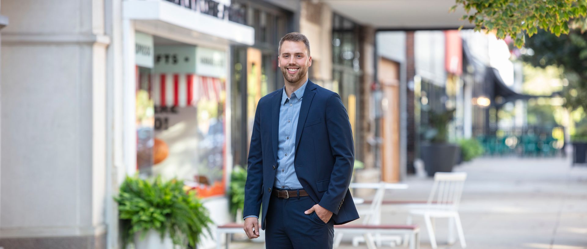 A smiling professional in a navy suit stands on an outdoor sidewalk lined with storefronts and plants.