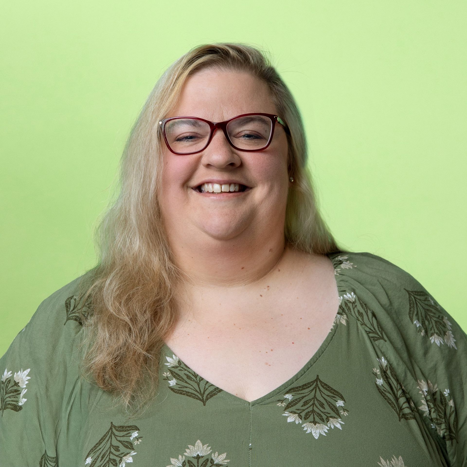 A smiling person with long blonde hair and red-rimmed glasses, wearing a patterned olive green top against a lime background.