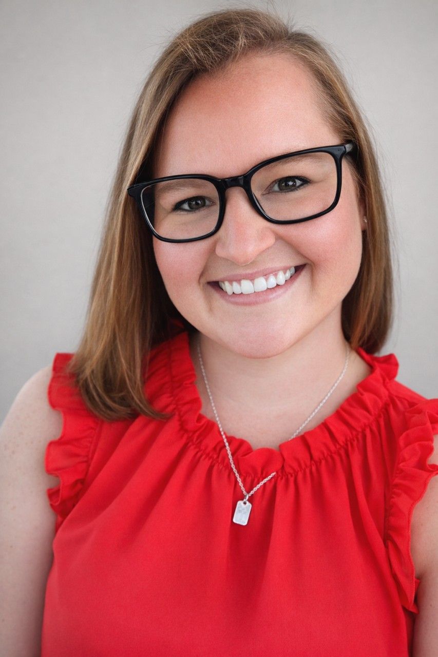 A smiling person with long light brown hair and black glasses, wearing a sleeveless red top and a silver necklace.