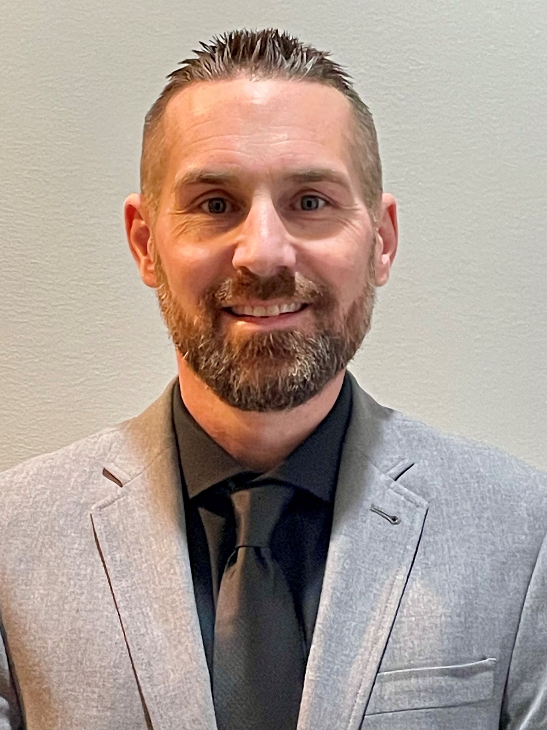 A person wearing a grey blazer, black collared shirt, and black tie, smiling against a neutral light-colored background.