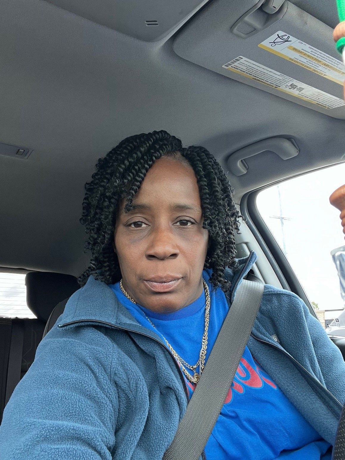 A person with dark, curled hair wearing a blue jacket and shirt, sitting in the driver's seat of a car.