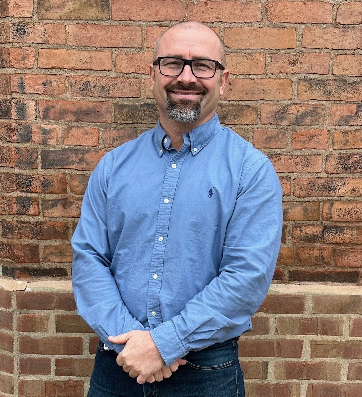 A person with glasses and a goatee wearing a blue button-down shirt, smiling in front of a brick wall.