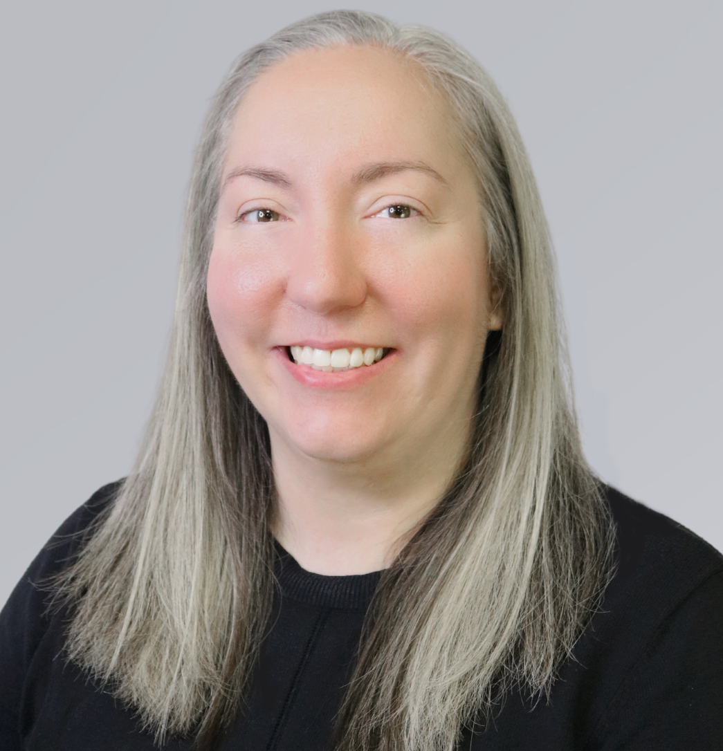A person with long, straight grey hair smiling at the camera against a plain, light grey background.