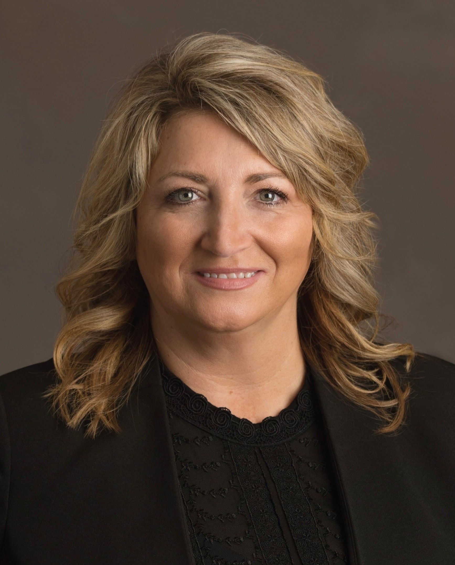 Headshot of a person with shoulder-length blonde hair, wearing a black blazer over a dark top against a neutral background.