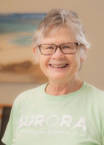 Smiling person with glasses wearing a light green Aurora t-shirt in a soft, bright indoor setting.
