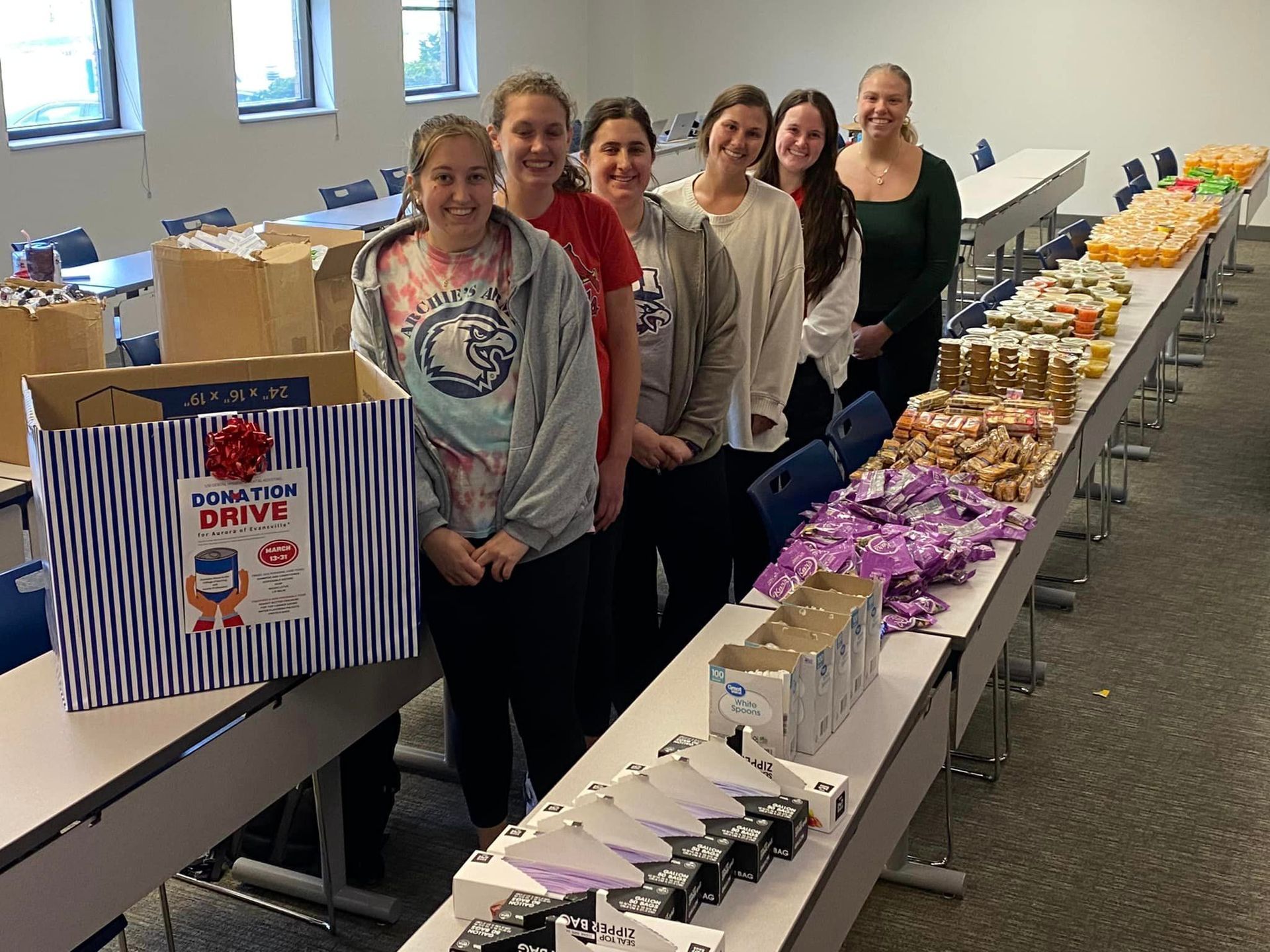 Six people stand behind a long table filled with organized food donations for a community drive.