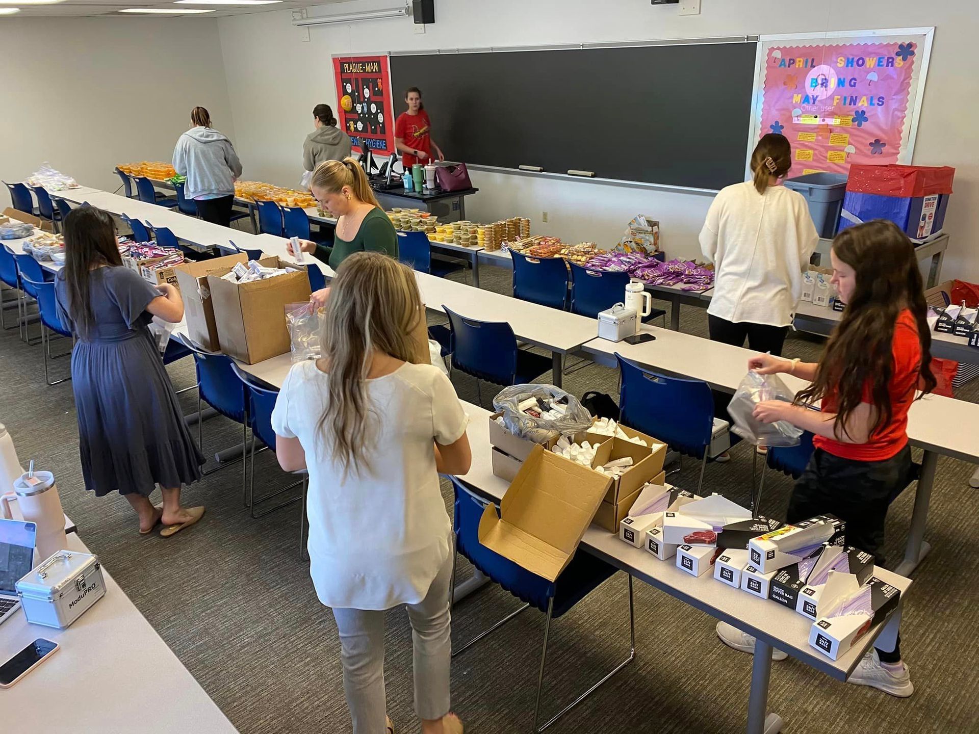 People pack school supplies and personal care items into boxes at long tables in a classroom.