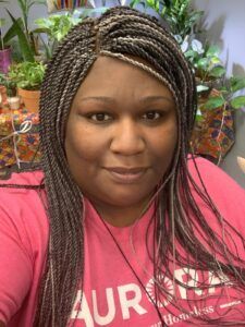 A person with braided hair and a soft smile wears a pink long-sleeved shirt in a room filled with potted plants.