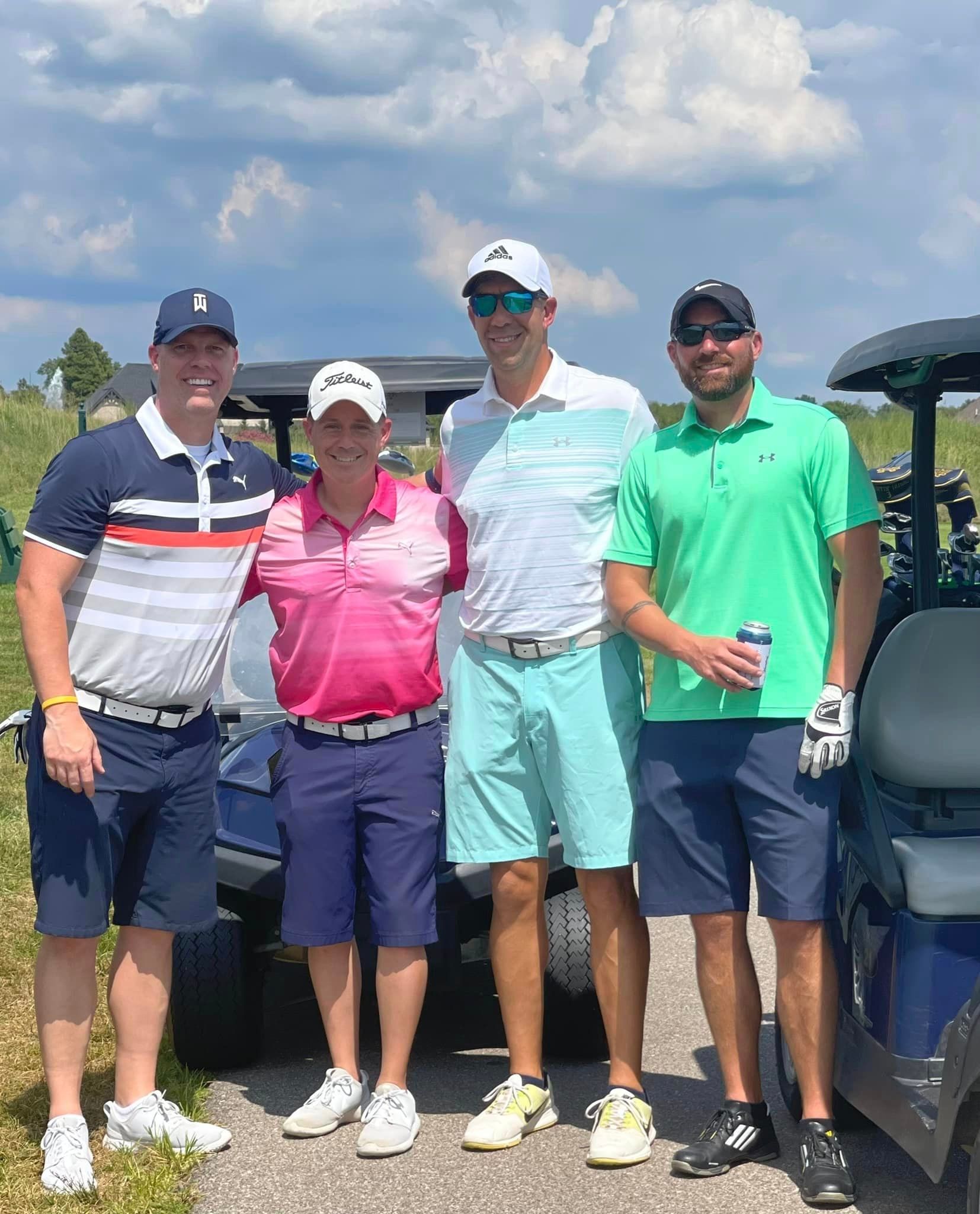 Four people standing in a row on a golf course, smiling and wearing golf attire with golf carts in the background.