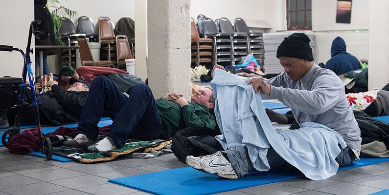 People rest on blue floor mats in a large indoor room, with one person sitting and folding a light-colored blanket.