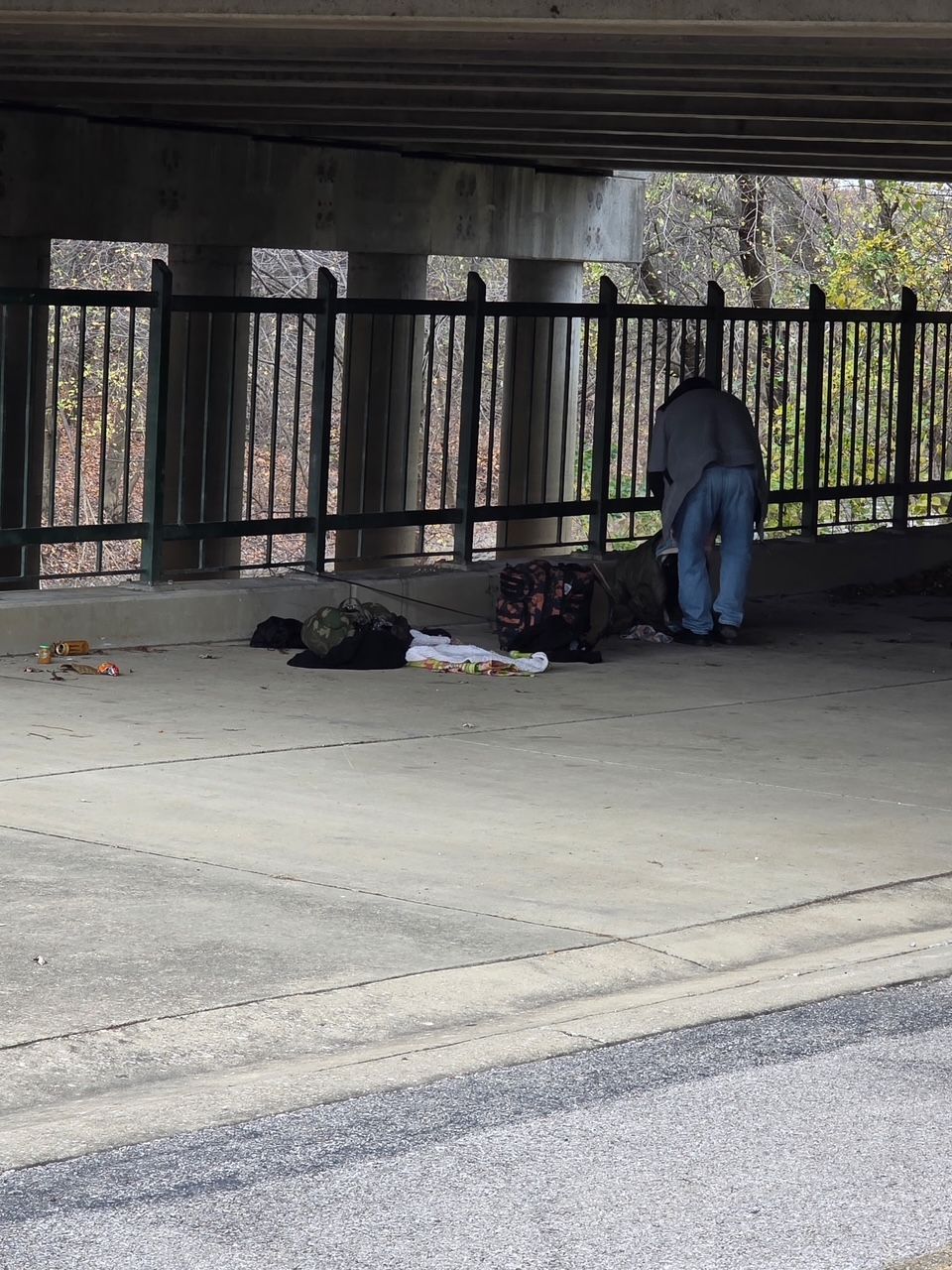 A person stands under a concrete overpass near a fenced area with belongings placed on the pavement nearby.