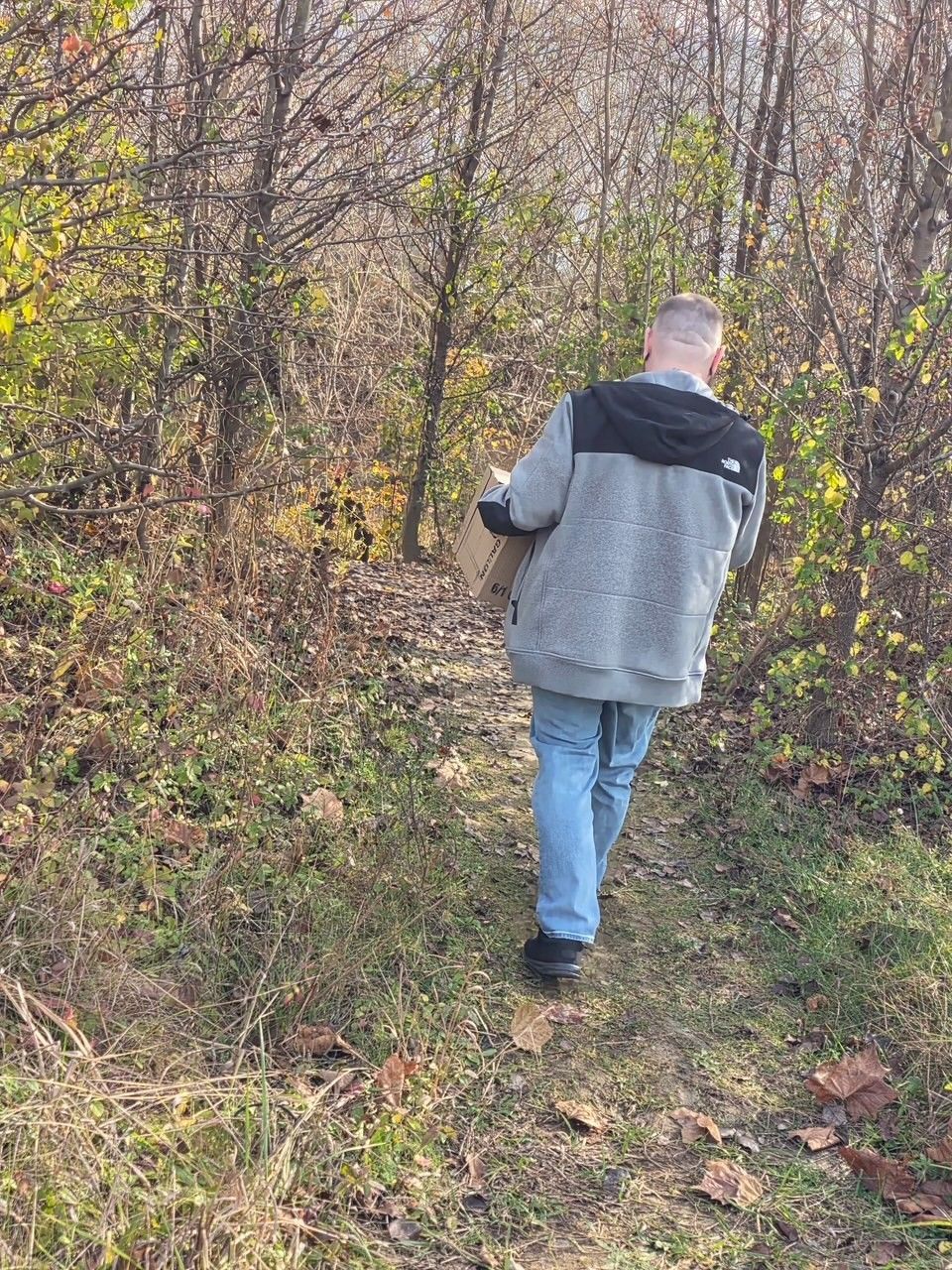 A person in a gray zip-up fleece and blue jeans carries a package while walking along a wooded, leaf-covered path.