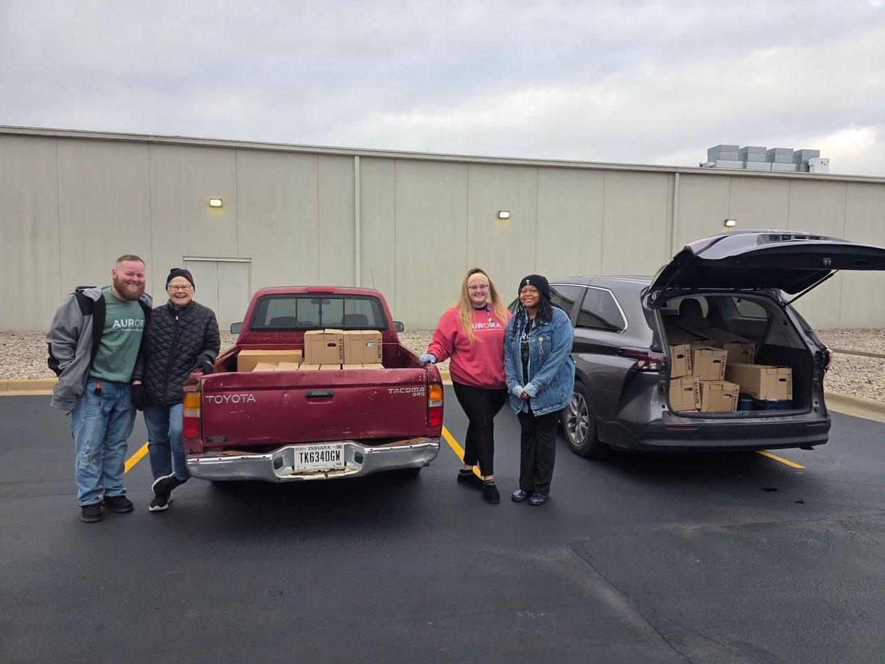 Four people stand by a red pickup truck and a grey SUV parked in a lot, both vehicles loaded with cardboard boxes.