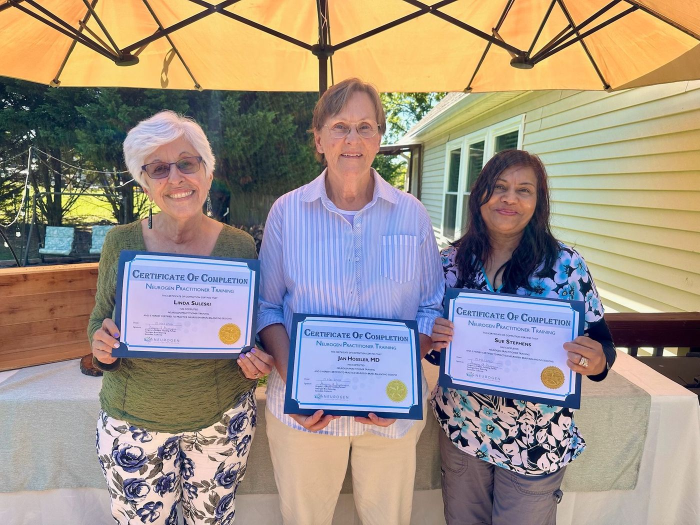 Three people holding certificates outdoors under a yellow patio umbrella