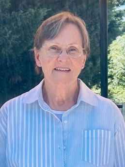 Smiling person wearing glasses and a light blue striped shirt outdoors in front of greenery