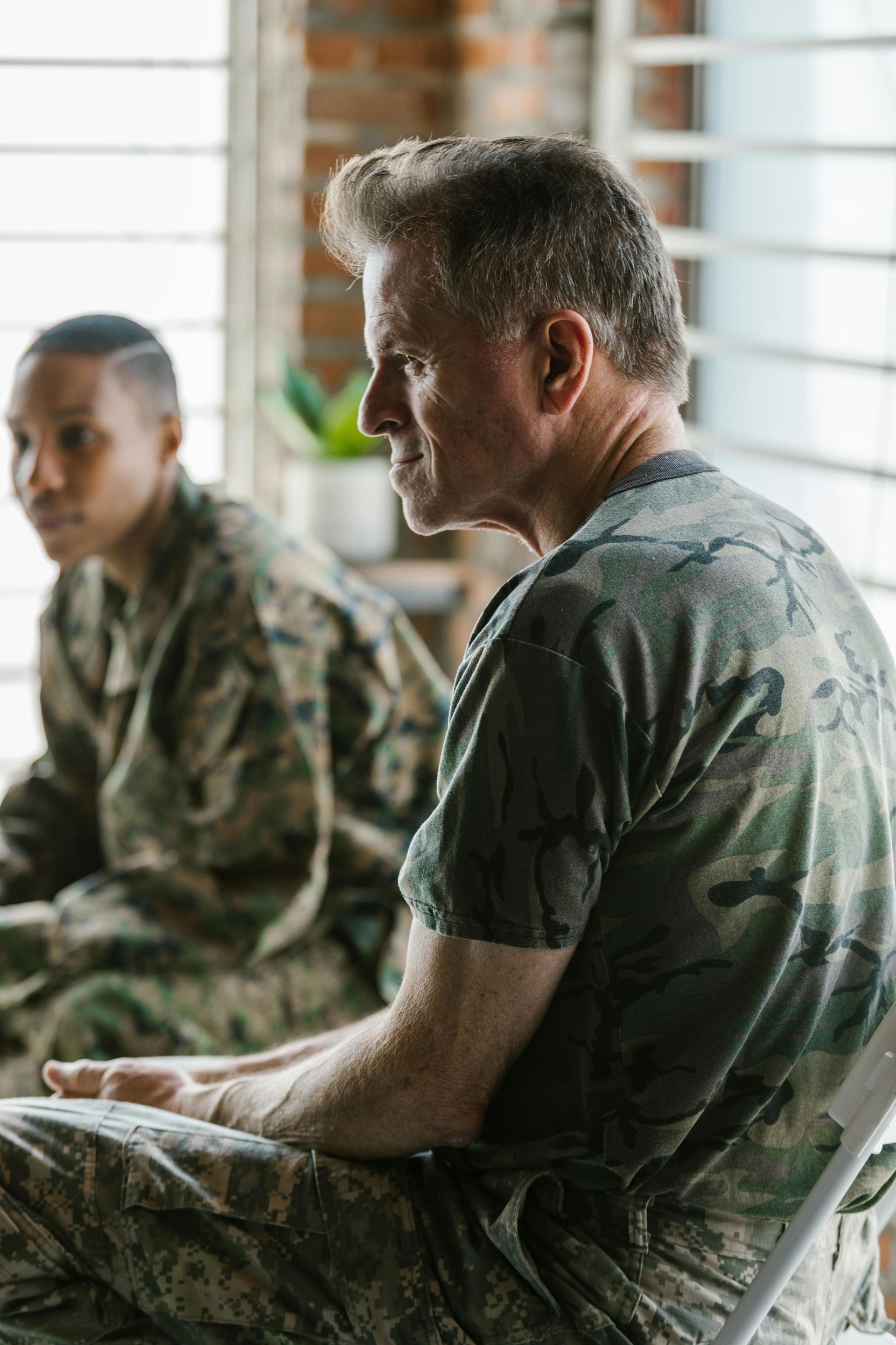 Two soldiers in camouflage uniforms sitting indoors by a window, one in profile, one blurred in background