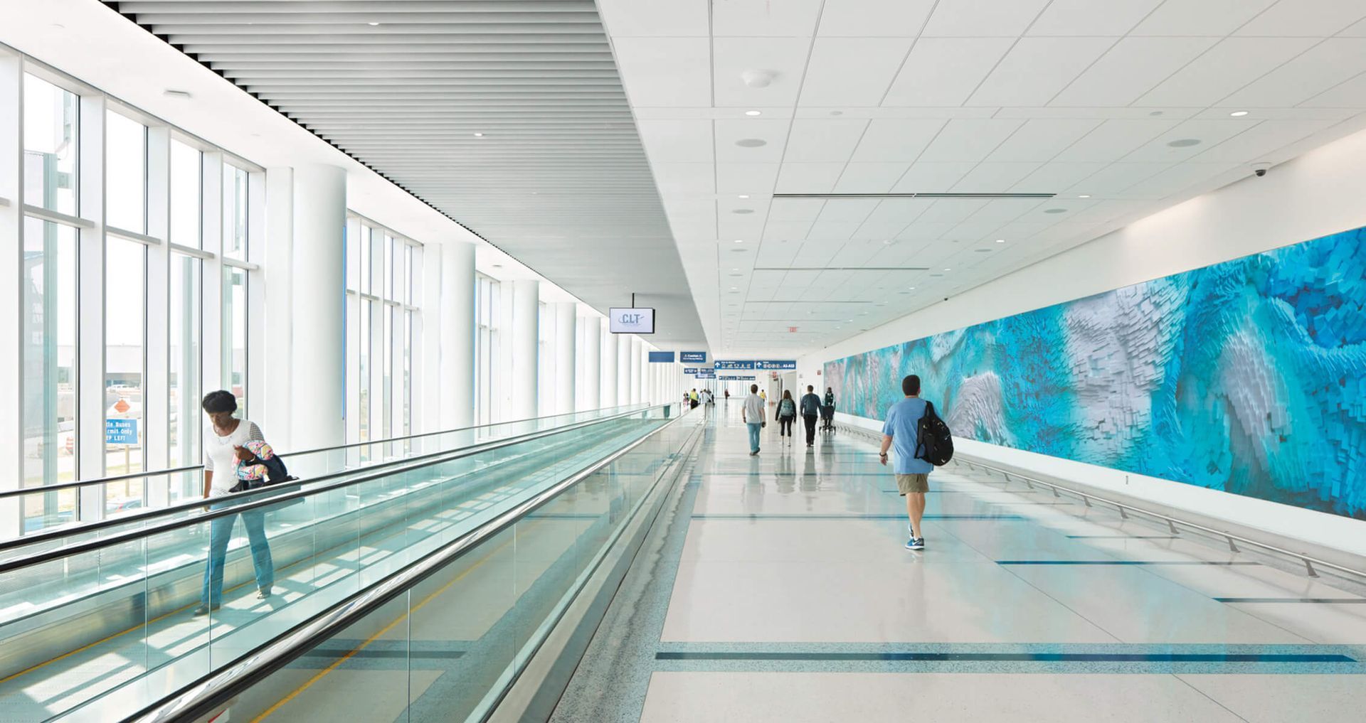 People walking on a moving walkway in a bright airport terminal with a blue art installation on a wall.