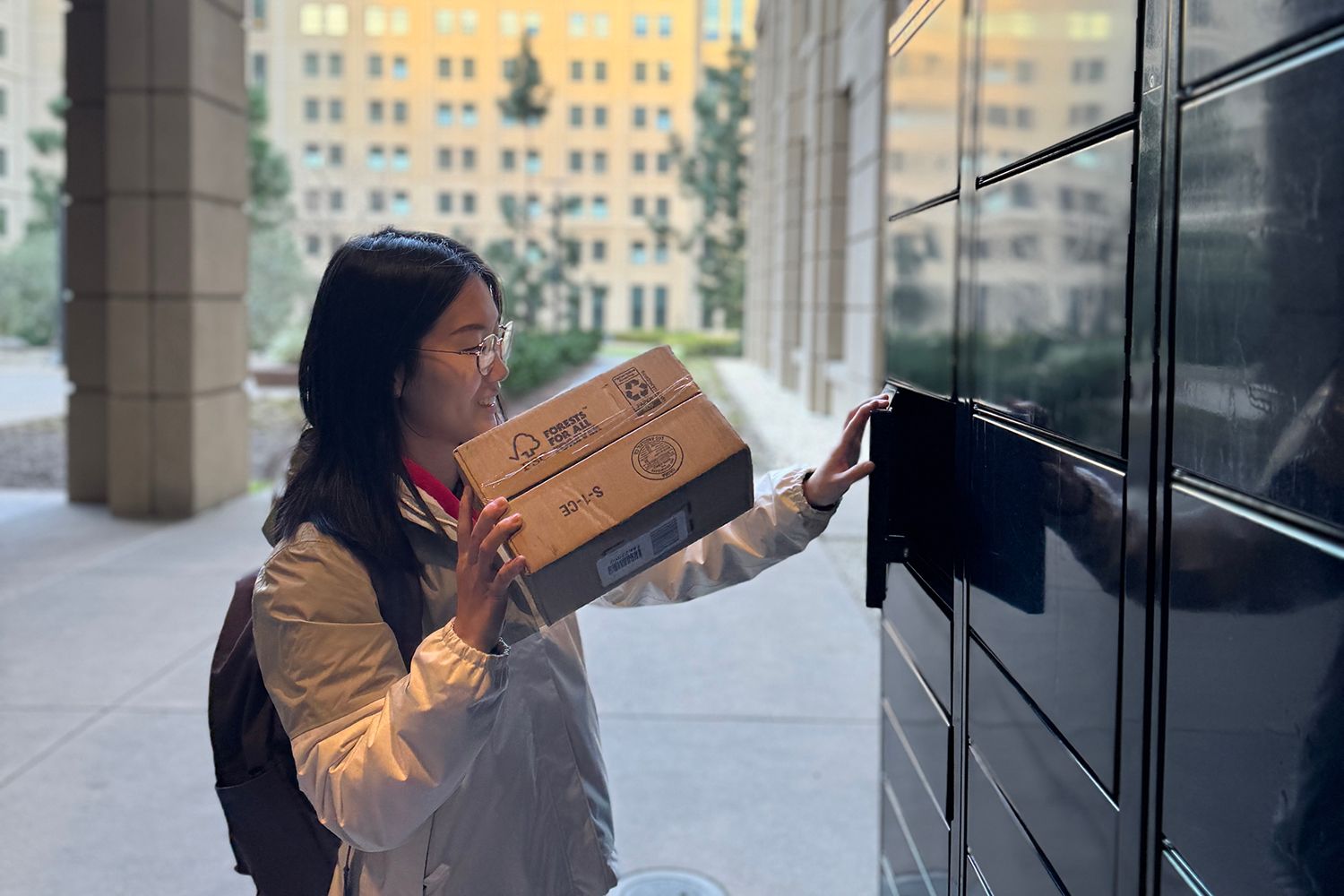 Woman depositing package in outdoor smart locker. Building and trees in the background.