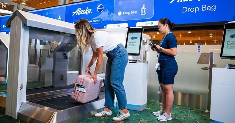 Woman putting a pink suitcase on a conveyor belt at an Alaska Airlines bag drop kiosk; employee nearby.