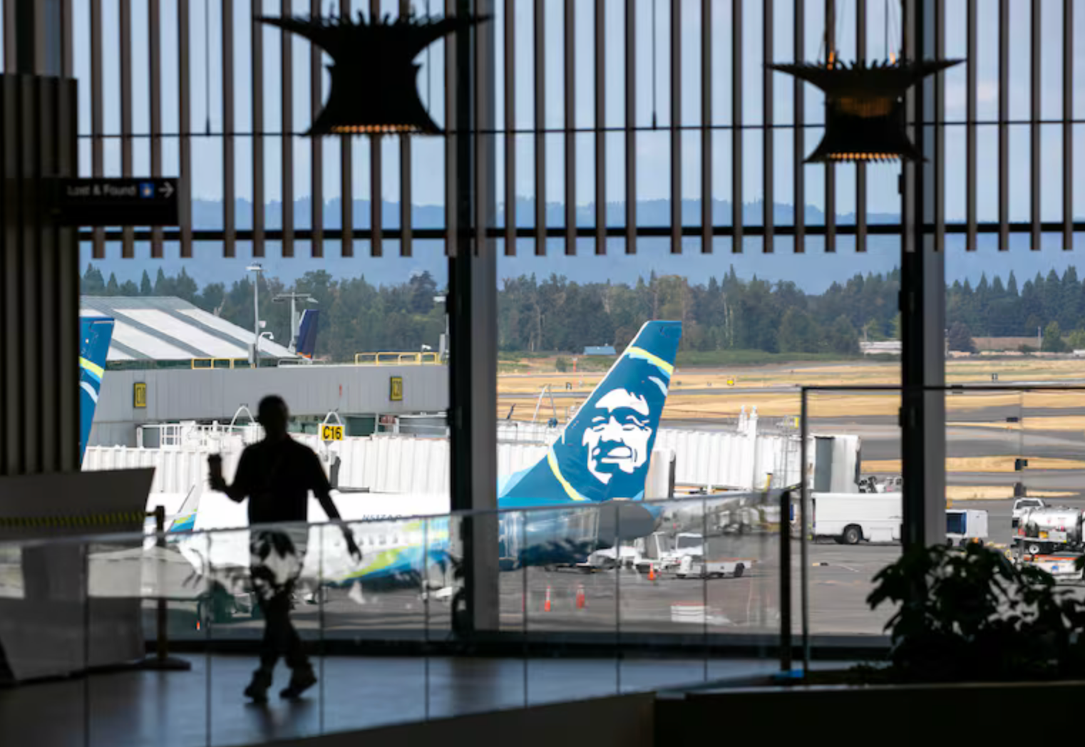 Silhouette of a person walking in an airport terminal, Alaska Airlines plane visible through windows, sunny day.