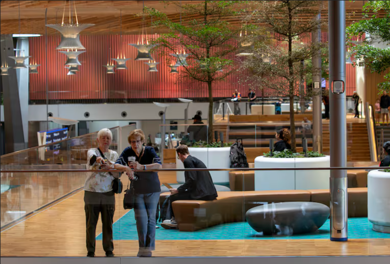 Two women stand near a glass railing, looking at an indoor seating area with trees, people, and modern lighting.