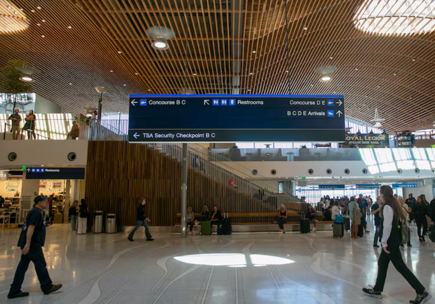Airport interior with signs directing passengers. People walk through a large, modern space with seating areas.