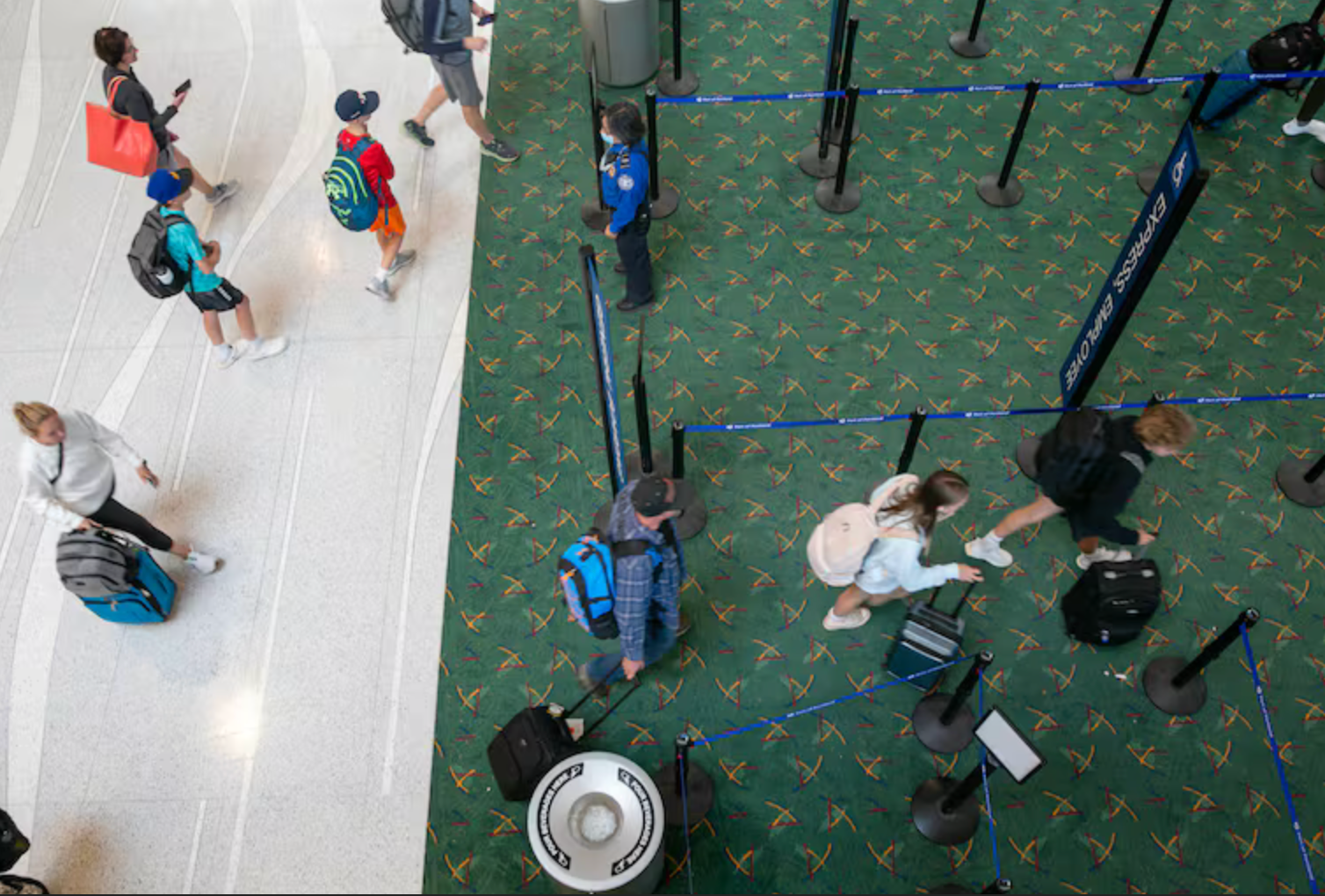 People in an airport. Some are walking, others waiting in line. Green carpet and white tile floor.