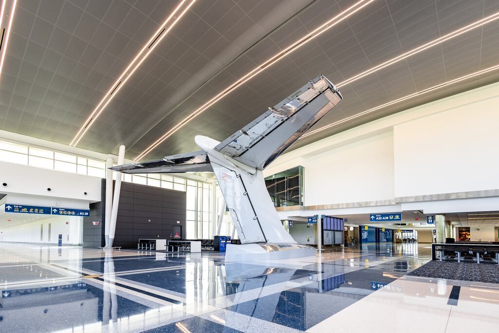 Aviation-themed airport interior with large aircraft wing sculpture in the center, reflective floor, and bright lighting.