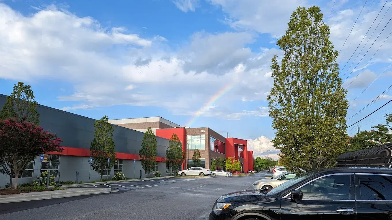 Modern red and gray building with cars parked in front, rainbow in the partly cloudy sky.