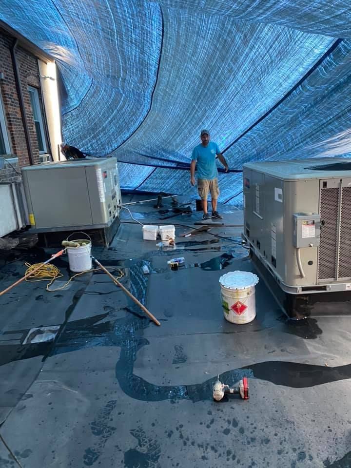 Person pressure-washes a rooftop under a blue tarp near HVAC units and cleaning buckets.