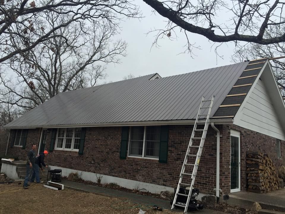Workers on a brick house roof with ladders, installing gray metal roofing on an overcast day