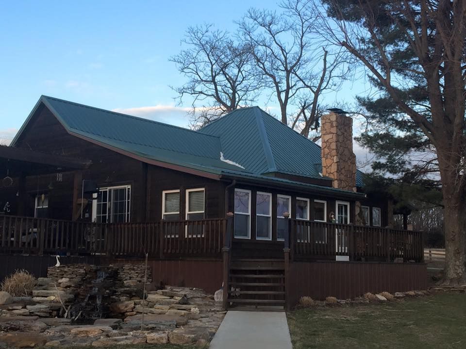 Dark wood cabin with green roof and stone chimney, set among bare trees on a hillside.