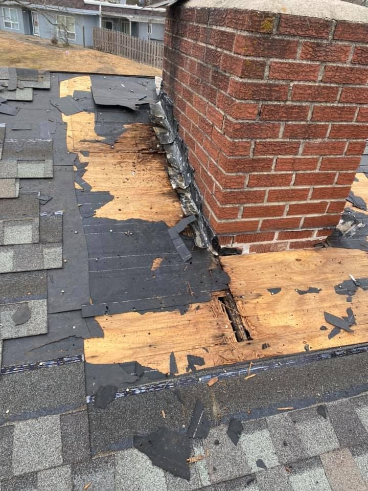 Damaged roof shingles and exposed plywood around a brick chimney