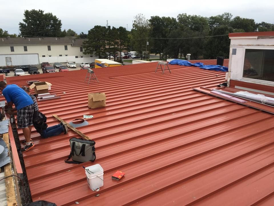 Workers installing a red metal roof on a building, with tools and materials spread across the rooftop.
