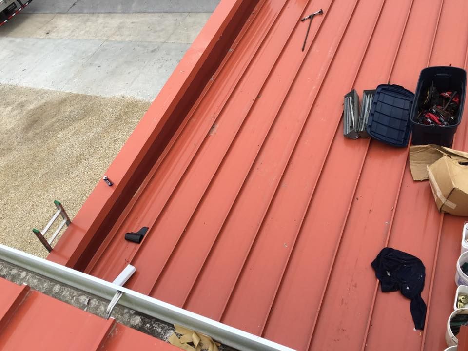 Tools and a blue rope spool on a red metal roof beside a ladder near a gravel surface
