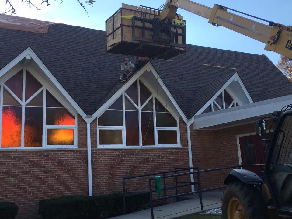 Worker on a lift repairing a brick church roof at dusk, with orange light glowing from the windows.