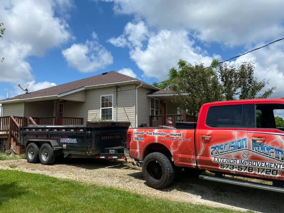 Red tow truck hauling a black trailer in front of a house under a partly cloudy sky