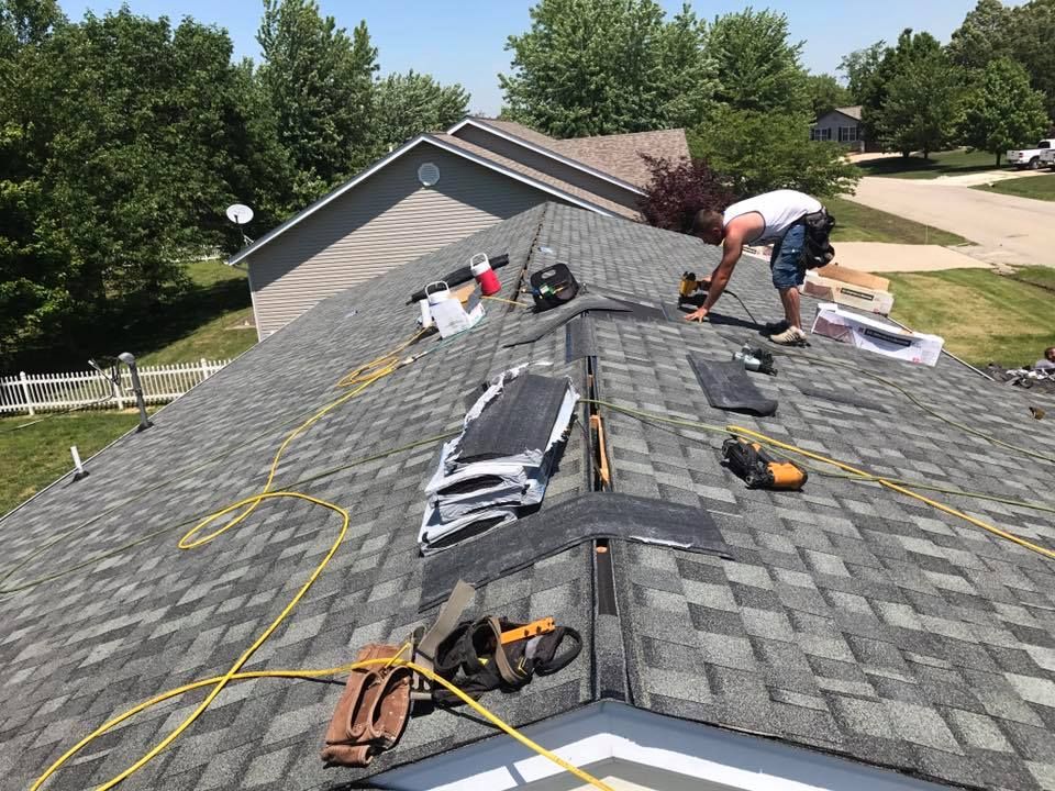 Workers repairing shingles on a steep gray roof with tools and materials spread out.