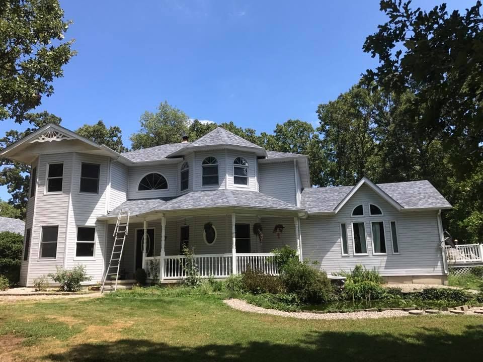 Large white two-story house with porch and gabled roof, set on a grassy lawn with trees and blue sky