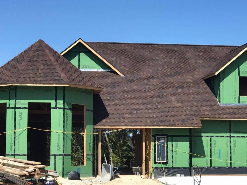 Green house under construction with dark shingle roof and stacked lumber in front