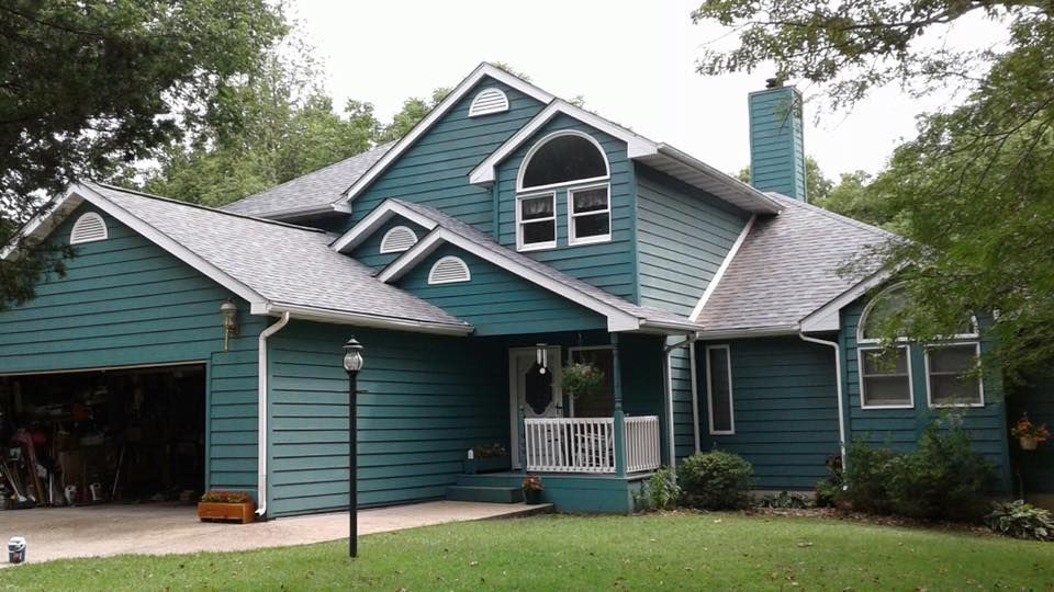 Teal house with white trim and gabled roof, front porch, and attached garage surrounded by trees