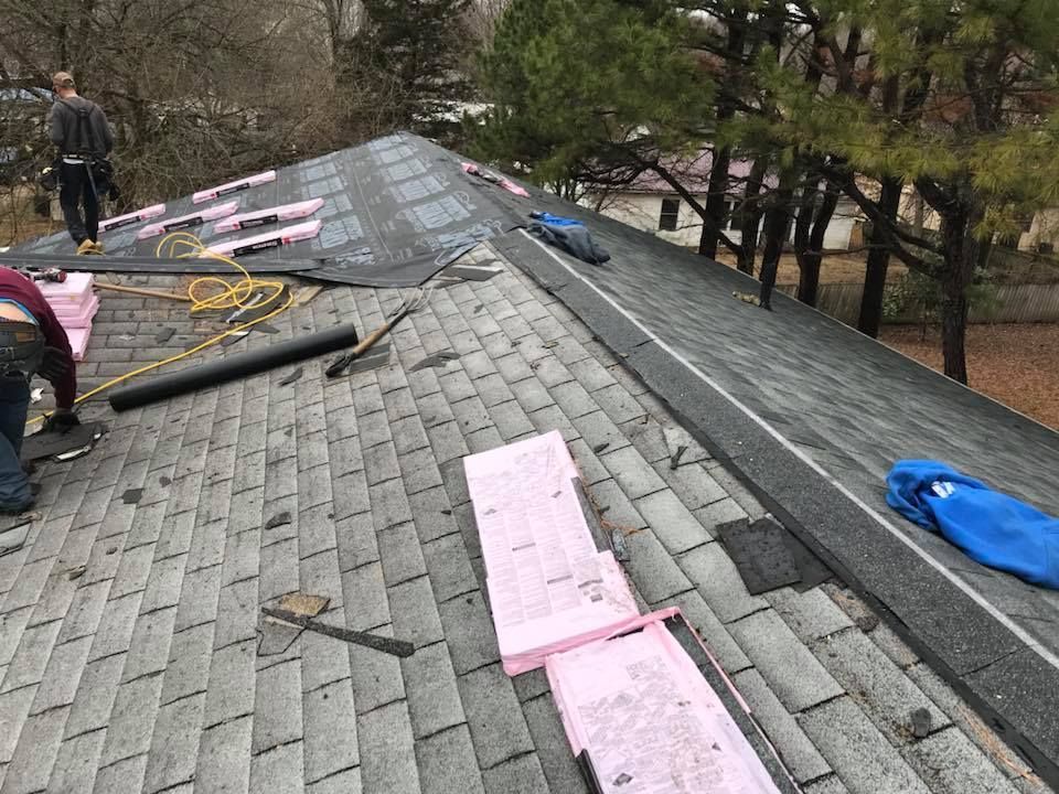 Roof workers installing pink underlayment on a gray shingled roof in a wooded area.