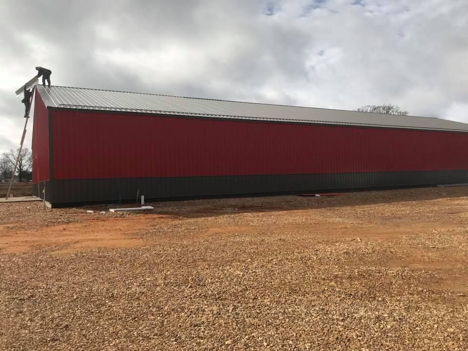 Long red metal barn with a gray roof on a gravel lot under a cloudy sky