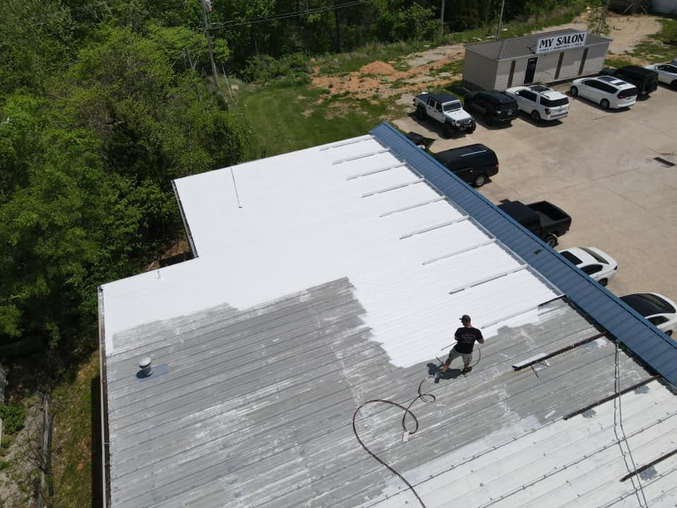 Worker spraying gray coating on a white flat roof near parked cars and trees
