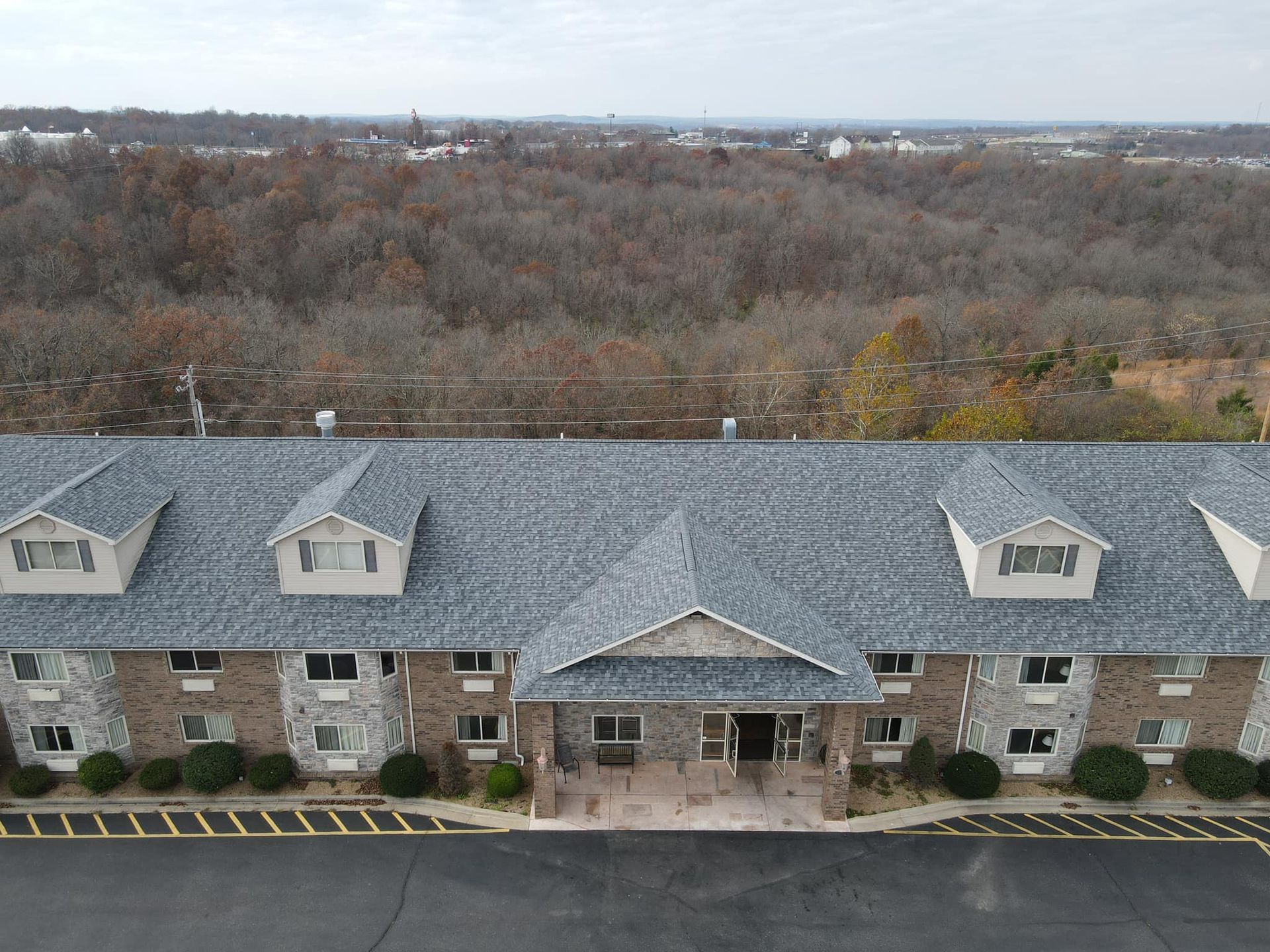 Aerial view of a stone apartment building with a gray roof, backed by leafless trees and a distant skyline.