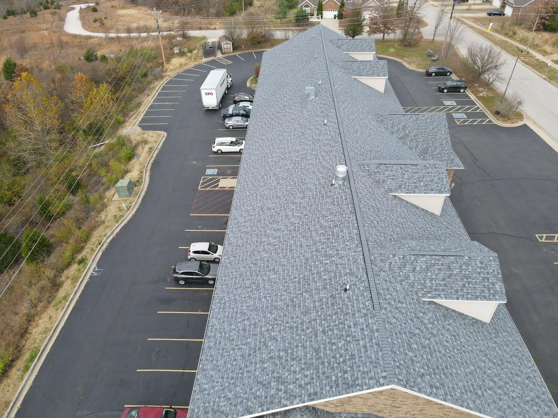 Aerial view of a long gray-roofed building with a parking lot and several parked vehicles.